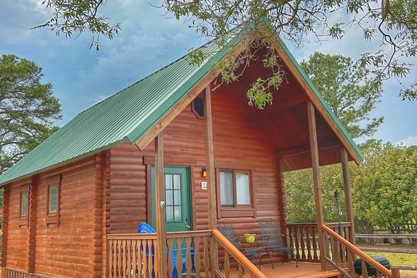 Red log cabin with green metal roof, front porch with wooden railings and steps, surrounded by trees and grass