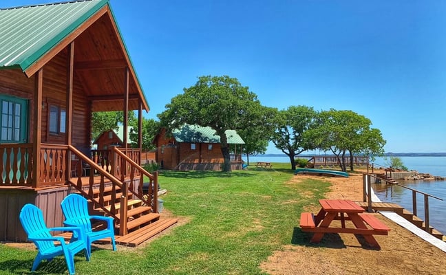 Waterfront cabin resort with red wooden buildings, blue Adirondack chairs, picnic tables, and lake view under clear sky